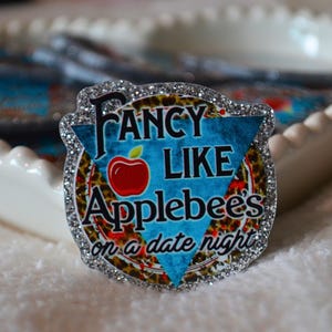 May include: A decorative coaster with a glittery border and the text "Fancy Like Applebees on a date night." The coaster features a red apple graphic and a blue and leopard print background. Several coasters are in a white, scalloped dish.
