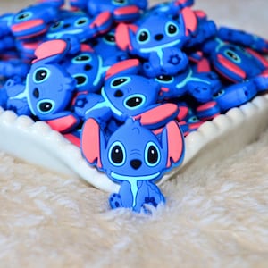 May include: A collection of blue and pink cartoon character charms. The charms feature a blue body, large black eyes, and pink ears. The charms are piled in a white, scalloped-edge dish, with one charm in focus.