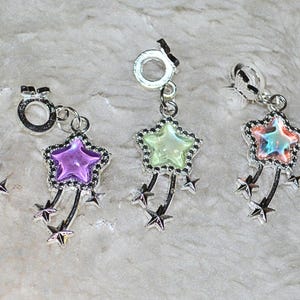 Puede incluir: Cinco colgantes de estrella plateados con centros coloridos y detalles de estrellas colgantes. Los colgantes presentan una variedad de colores, incluyendo amarillo, morado, verde y una mezcla de rosa y azul. Cada colgante tiene un pequeño lazo.
