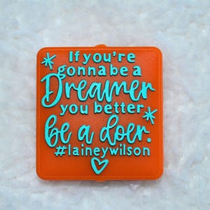 Op de afbeelding: Oranje vierkant met turquoise tekst: "If you're gonna be a Dreamer you better be a doer. #laineywilson", met ster- en hartaccenten. Motiverende quote.