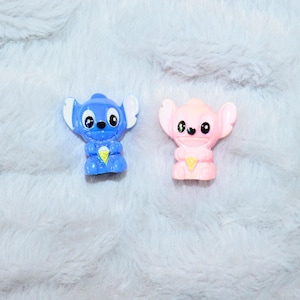 May include: Two small, plastic figurines of the character Stitch, one blue and one pink, are displayed. The blue Stitch has white ears and a yellow ice cream cone. The pink Stitch has white ears and a yellow ice cream cone. The figurines are on a soft, gray surface.