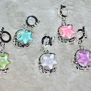 Puede incluir: Cinco colgantes plateados con diseños de estrellas y lunas crecientes de colores. Cada colgante presenta una estrella de diferente color: verde, azul, rosa, blanco y morado. Los colgantes son probablemente para pulseras o collares.