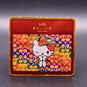 May include: A square Coach wallet featuring a dark red leather top section with a gold embossed logo. The lower portion displays a vibrant, multi-colored repeating pattern with a central graphic of a white Hello Kitty character wearing a colorful bow.