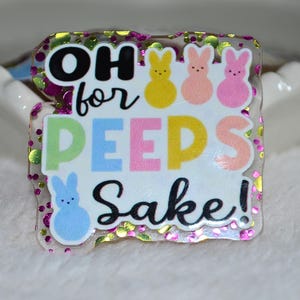 以下が含まれることがあります： 「OH for PEEPS Sake!」というフレーズが黒、パステル、黄色の文字で書かれた、キラキラした縁取りの透明なバッジ。黄色、ピンク、青色の漫画のウサギが描かれています。背景は白です。