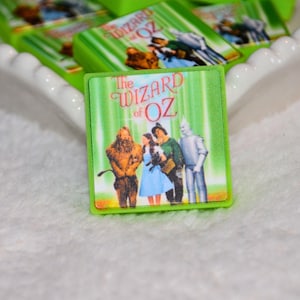 May include: Green square-shaped magnets featuring the iconic characters from "The Wizard of Oz" movie. The magnets have a green border and a vertical striped background. The text "The Wizard of Oz" is displayed on the front.
