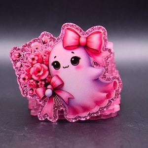 May include: A pink ghost-shaped acrylic charm featuring a glittery border, a large pink bow on its head, and a bouquet of pink flowers held by a matching pink ribbon. The ghost has large black eyes, rosy cheeks, and a small mouth, with a layered base.