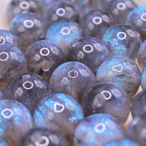 May include: A collection of round, translucent beads featuring a dark gray base with swirling patterns and vibrant blue glitter inclusions. Each bead has a smooth, polished surface and a central hole, creating a reflective and textured appearance.