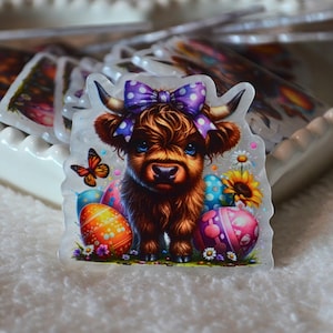 May include: A clear sticker depicting a Highland cow with a purple bow, Easter eggs, a butterfly, and flowers. The cow has brown fur and blue eyes. The sticker is in a heart-shaped dish with other stickers.
