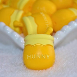 May include: A yellow honey pot with the word "HUNNY" in white lettering. The pot has a yellow lid and a small dipper. Other yellow honey pots are in the background, all on a white surface.