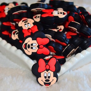 Könnte beinhalten: Eine Sammlung von schwarzen, roten und weißen Minnie-Mouse-förmigen Charms. Die Charms zeigen Minnie's Gesicht, eine rote Schleife und einen schwarzen Körper. Sie sind in einer weißen Schale mit gewelltem Rand aufgestapelt.
