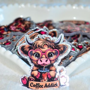Pode incluir: Um adesivo brilhante com uma vaca de desenho animado com óculos em forma de coração, bebendo café. A vaca é marrom e branca, com nariz e orelhas rosas. O texto "Coffee Addict" está abaixo.