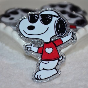 May include: A Snoopy-themed pin featuring the cartoon dog wearing sunglasses and a red shirt with a white heart. The pin has a glittery silver border and is set against a white background. Additional Snoopy pins are visible in the background.