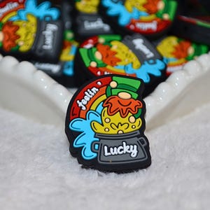 Puede incluir: Una colección de coloridos dijes para zapatos con temática de duende. Los dijes tienen una base negra con una olla de oro, un arcoíris y la palabra "Lucky". Otros dijes son visibles en el fondo.