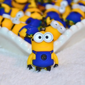Op de afbeelding: Een verzameling gele en blauwe Minion-beeldjes, met één in scherpe focus. De beeldjes hebben blauwe overalls, zwarte schoenen en grote, ronde ogen. Ze zijn gerangschikt in een wit, geschulpte schaal, wat een speels thema suggereert.
