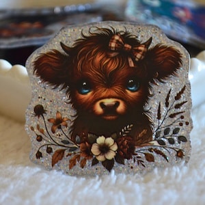 Pode incluir: Um adesivo brilhante com uma vaca Highland com pelo castanho, olhos azuis e um laço xadrez. A vaca é rodeada por detalhes florais e de folhas em tons de castanho e creme. O adesivo tem um fundo brilhante e texturizado.