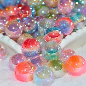 May include: A collection of round, translucent beads featuring an iridescent finish with embedded glitter particles. The beads display a vibrant spectrum of colors including pink, blue, orange, and green, creating a shimmering, reflective surface.