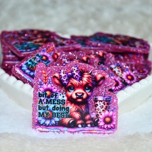 May include: A collection of glittery pink stickers featuring a cartoon Highland cow with a bow, sitting among flowers and a leopard print cup. The text on the sticker reads "bit of A MESS but doing MY BEST".