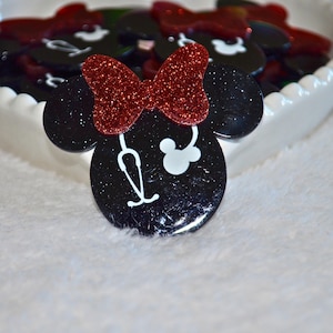 Può includere: Abbellimenti neri a forma di Minnie Mouse con fiocchi rossi glitterati e dettagli bianchi. Un design di stetoscopio bianco è sul viso della forma di Minnie Mouse. Gli abbellimenti sono in un piatto bianco a forma di cuore.