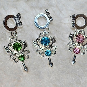 May include: Five silver-toned charm pendants with decorative designs and colorful gemstones. Each charm features a heart-shaped design with wings, a gemstone, and a dangling element. The gemstones are in various colors, including clear, green, blue, pink, and purple.