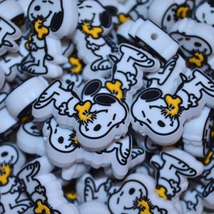Pode incluir: Uma cole&ccedil;&atilde;o de contas brancas em forma de Snoopy, com contornos pretos e detalhes amarelos. As contas est&atilde;o empilhadas, mostrando a personagem de desenho animado em v&aacute;rias poses. Estas contas s&atilde;o provavelmente para artesanato ou para fazer joias.