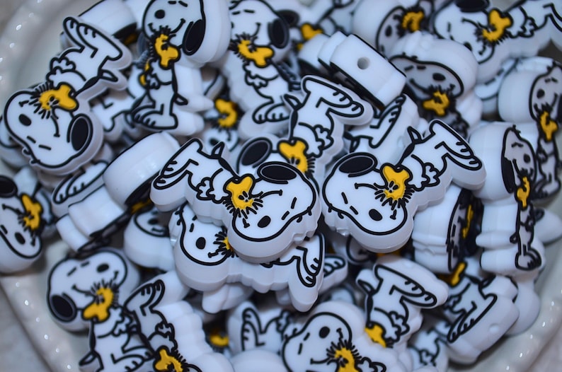 Pode incluir: Uma cole&ccedil;&atilde;o de contas brancas em forma de Snoopy, com contornos pretos e detalhes amarelos. As contas est&atilde;o empilhadas, mostrando a personagem de desenho animado em v&aacute;rias poses. Estas contas s&atilde;o provavelmente para artesanato ou para fazer joias.
