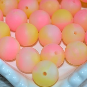 May include: Close-up of a collection of pink and yellow ombre beads. The beads are round with a hole through the center, and are arranged on a white, scalloped-edge tray. The beads have a smooth, matte finish.