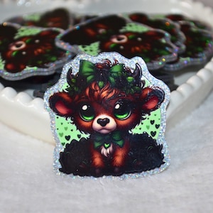Pode incluir: Uma coleção de autocolantes com uma vaca Highland de desenho animado com uma gravata borboleta verde e um laço verde na cabeça. A vaca tem olhos verdes grandes e uma borda brilhante. Os autocolantes estão numa tigela branca em forma de coração.