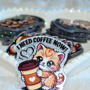 May include: A glittery sticker featuring a cartoon kitten holding a coffee cup. The kitten is orange and white with large blue eyes. The coffee cup is tan with a brown lid and a white heart. Text above the kitten reads "I NEED COFFEE NOW!"