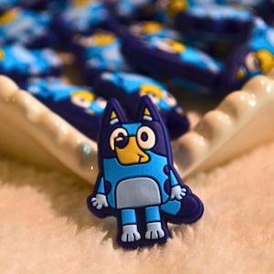 Op de afbeelding: Een verzameling blauwe en lichtblauwe cartoonbedels. De bedels hebben de vorm van een hond met een lichtblauw lichaam, donkerblauwe accenten en een gele snavel. De bedels liggen op een wit dienblad.