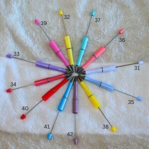 May include: A collection of colorful, small tools with long, thin metal shafts and various colored handles and tips. The tools are arranged in a circular pattern, with each tool radiating outwards from a central point. The colors include pink, yellow, blue, and purple.
