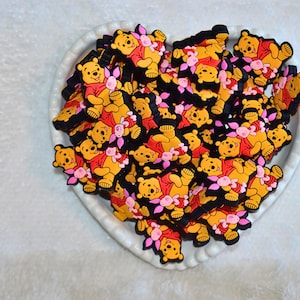May include: A heart-shaped dish filled with numerous yellow, red, and black Winnie the Pooh character charms. Each charm features the bear with a pink flower. The charms are on a white, textured surface.