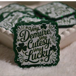 May include: Green and white glittery rectangular pin with the words "Very Demure, Very Cutesy, Very Lucky" and shamrock designs. Several similar pins are in a white heart-shaped dish.