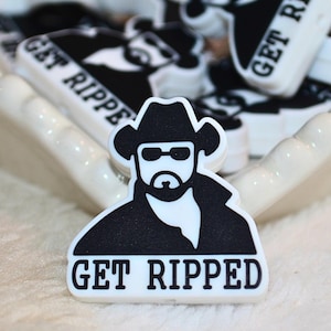 May include: A black and white shoe charm featuring a cowboy with sunglasses and the text "GET RIPPED". The charm is a rectangular shape with a white background and black details. Other similar charms are visible in the background.