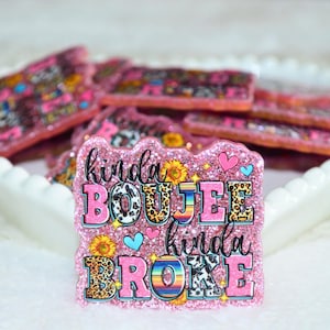 May include: Pink glittery badge with the text "kinda BOUJEE kinda BROKE". The letters have various patterns, including leopard print, cow print, and a rainbow stripe. The badge is decorated with sunflowers and hearts.