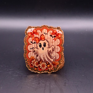 Puede incluir: Un broche rectangular de acrílico con una ilustración central de un fantasma blanco con un lazo rojo en la cabeza. El fantasma está rodeado de lazos naranjas, pequeños murciélagos negros y flores de otoño. El borde tiene un acabado festoneado dorado.