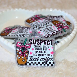 Könnte beinhalten: Glitzernde Acrylstifte mit dem Text "SUSPECT" und "Iced coffee". Die Stifte zeigen einen Cartoon-Eiskaffee mit einer rosa Schleife und dem Satz "CLAIMS SHE'S BROKE BUT SPENDS ALL HER MONEY ON Iced coffee."