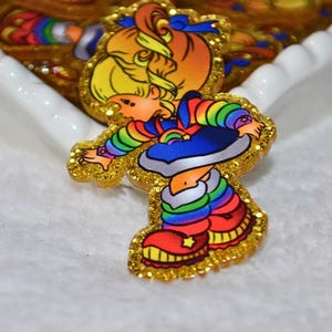 Puede incluir: Un pin colorido y brillante con un personaje de dibujos animados con un atuendo de arcoíris, sombrero azul y zapatos rojos. El pin tiene un borde dorado brillante. El diseño recuerda a una caricatura vintage.