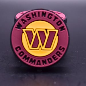 May include: A circular shoe charm featuring the Washington Commanders logo. The design includes a burgundy background with a yellow circle in the centre, a burgundy letter W, and the text WASHINGTON COMMANDERS in black lettering around the edge.