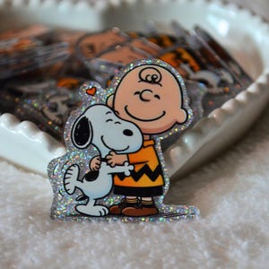 May include: A glittery sticker featuring Charlie Brown hugging Snoopy. Charlie Brown is wearing an orange shirt with a black zig-zag pattern and black shorts. Snoopy is white with black ears and spots. A small heart is above Snoopy's head.