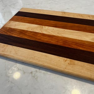 Puede incluir: Una tabla de cortar rectangular de madera con rayas alternas de madera clara y oscura. La tabla presenta una variedad de tonos de madera, incluyendo beige claro, marrón medio y marrón oscuro. Un pequeño logotipo es visible en la esquina.