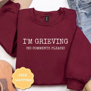 May include: A maroon long-sleeve crewneck sweatshirt featuring white text printed on the chest that reads I'M GRIEVING (NO COMMENTS PLEASE). The garment has a ribbed collar, cuffs, and hem, with a black brand label visible at the inner neckline.