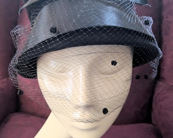 May include: A black hat with a veil, bow, and decorative dots. The hat is displayed on a mannequin head. The veil is a fine mesh that covers the face. The hat has a rounded crown and a brim.