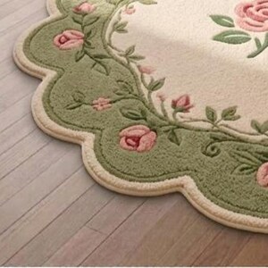 Op de afbeelding: Een vloerkleed met een geschulpte rand, een cr&egrave;mekleurig midden en een brede saliegroene rand. De rand is versierd met een herhalend patroon van roze rozenbloesems, knoppen en groene bladeren, met extra rozen in het midden.