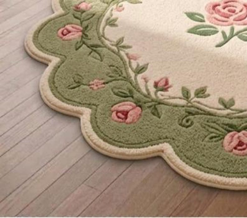 Op de afbeelding: Een vloerkleed met een geschulpte rand, een cr&egrave;mekleurig midden en een brede saliegroene rand. De rand is versierd met een herhalend patroon van roze rozenbloesems, knoppen en groene bladeren, met extra rozen in het midden.
