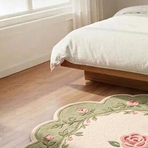 Op de afbeelding: Een vloerkleed met geschulpte randen, voorzien van een cr&egrave;mekleurig midden met een groot roze rozenmotief, omringd door een saliegroene rand versierd met kleinere roze rozenbloesems en groene bladranken, geplaatst op een lichte houten vloer.