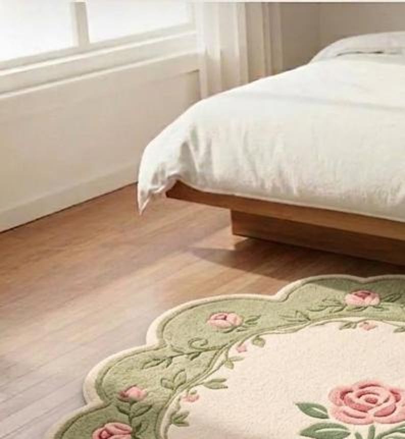 Op de afbeelding: Een vloerkleed met geschulpte randen, voorzien van een cr&egrave;mekleurig midden met een groot roze rozenmotief, omringd door een saliegroene rand versierd met kleinere roze rozenbloesems en groene bladranken, geplaatst op een lichte houten vloer.
