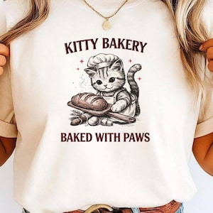 以下が含まれることがあります： クリーム色のTシャツ。コック帽をかぶった猫がパンを持っているグラフィック。テキスト「KITTY BAKERY」と「BAKED WITH PAWS」が猫のイラストの上と下に印刷されています。