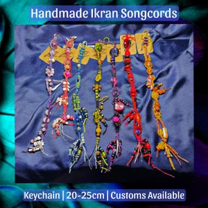 May include: Six handmade Ikran Songcords, each 20-25cm long, featuring varied colourful beads, knotted cords, and charms. The collection includes purple, red, blue, yellow, and orange designs, each attached to a small brown tag on a blue fabric.