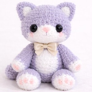 May include: A handmade crochet cat doll in lavender, white, and pink. The cat has large black eyes, a pink nose, and a cream-colored bow tie. The paws and inner ears are white with pink accents. A charming, soft toy.