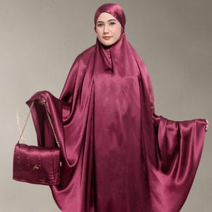 May include: A burgundy satin prayer outfit, comprising a head covering and a flowing garment. A matching burgundy purse with a gold chain strap is held. The outfit has a smooth, lustrous appearance.